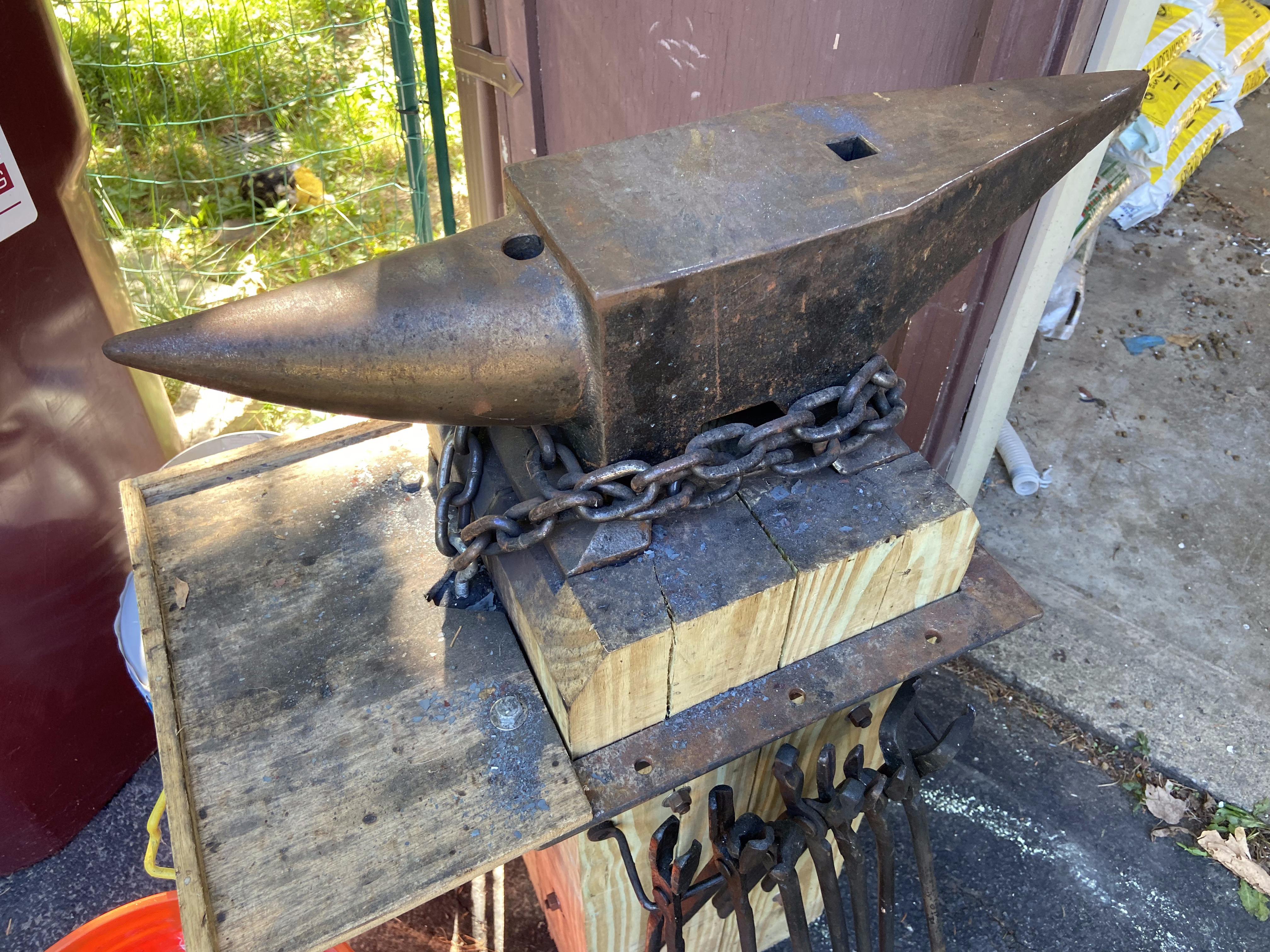 Heavy cast steel anvil chain-damped to a hardwood stump, tongs and tools arranged below