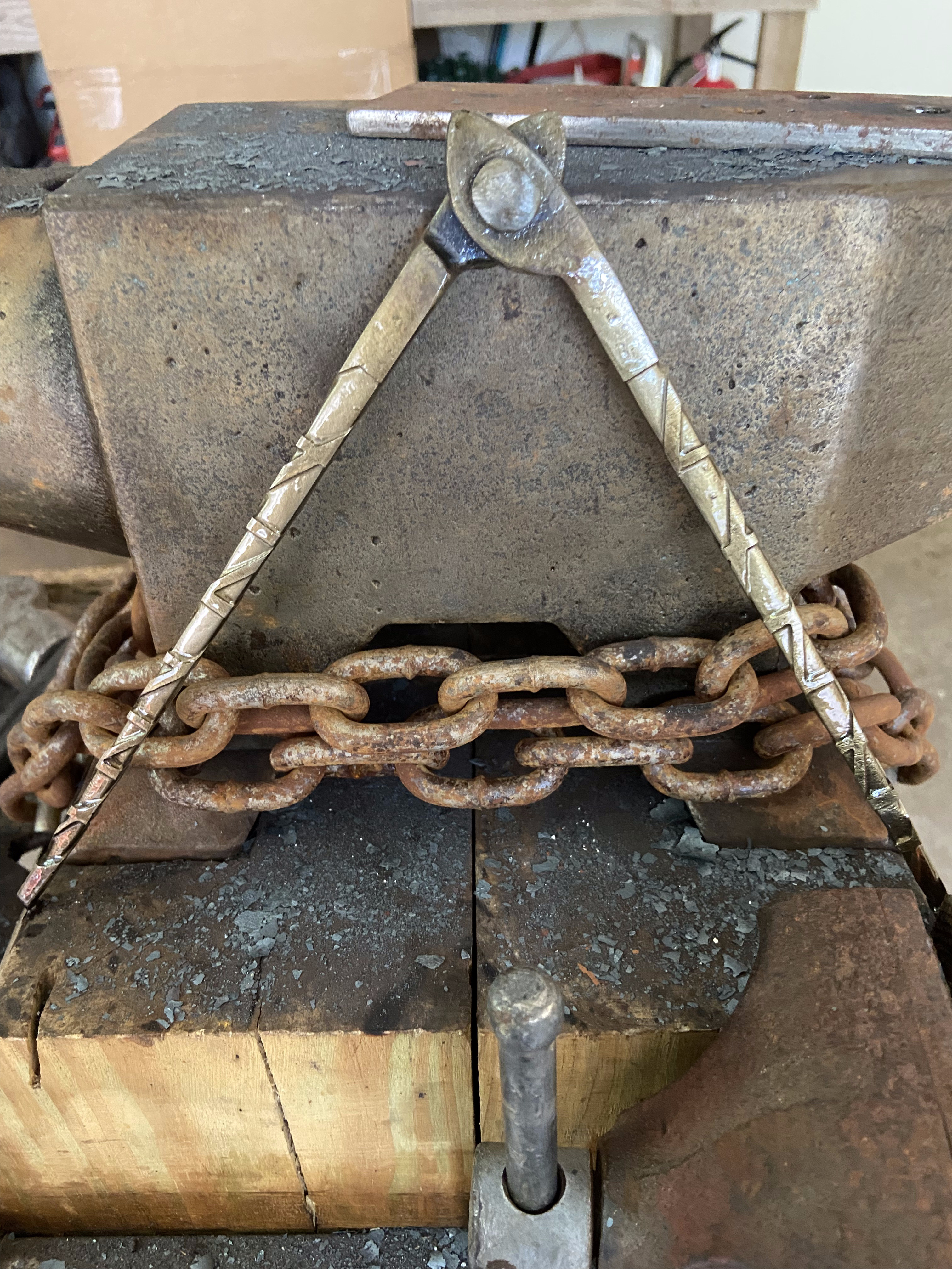 Forged compass dividers standing upright on the anvil face, chain-wrapped stump in the background