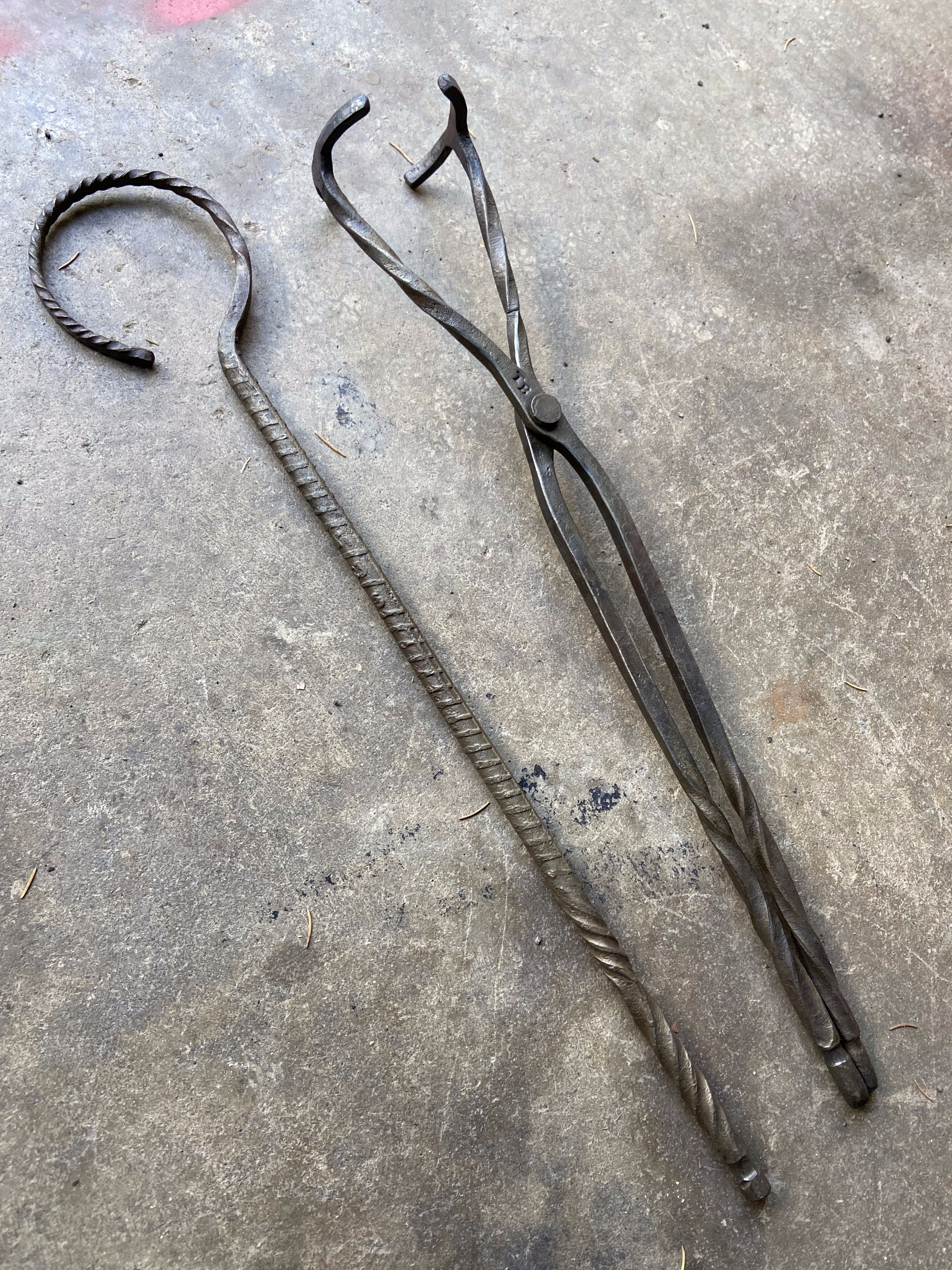 Forged fireplace hook with twisted handle and crucible tongs laid flat on concrete — two finished pieces