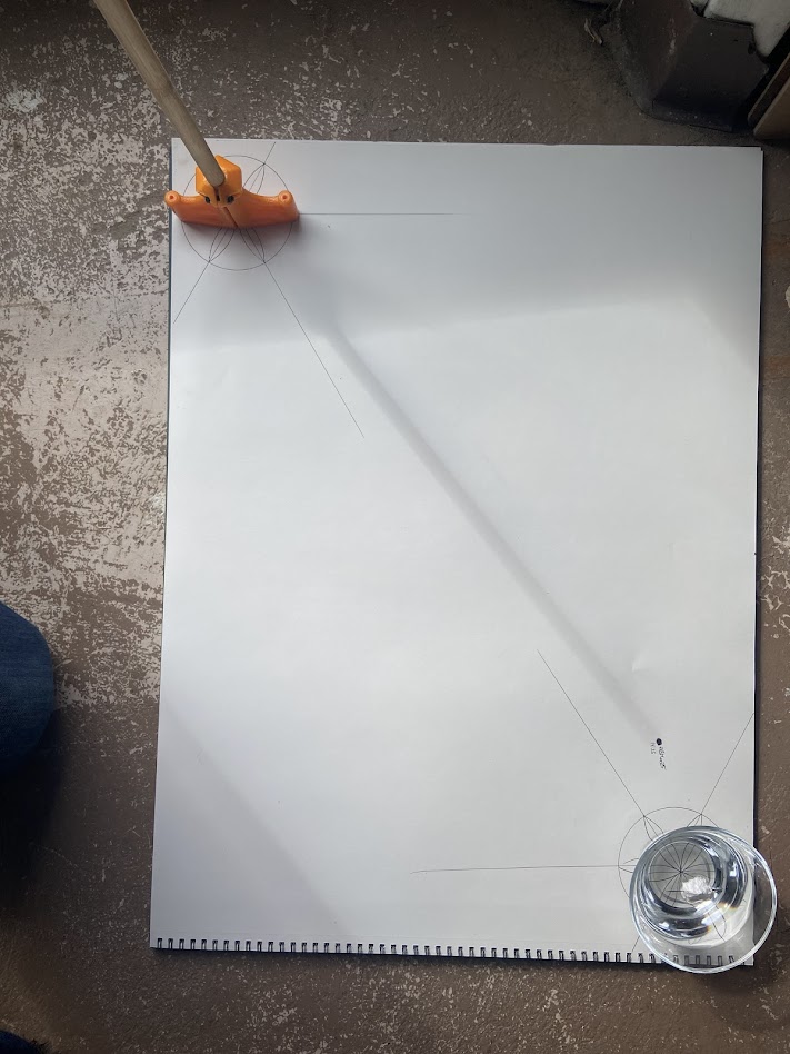 Gnomon shadow traced diagonally across a large white paper on a concrete floor at solar noon