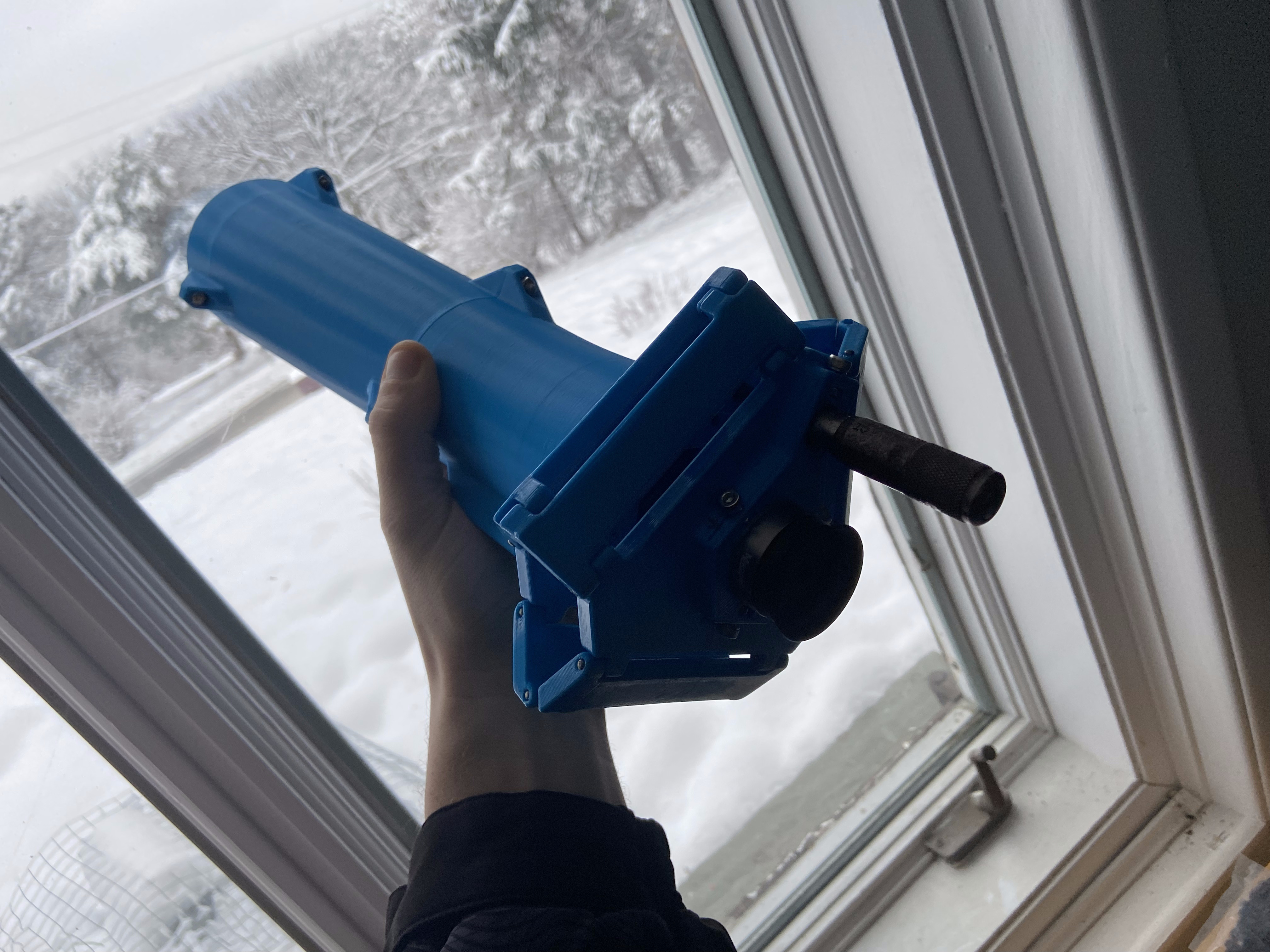 Blue 3D-printed telescope tube held up at a snowy window — focuser and mirror cell visible
