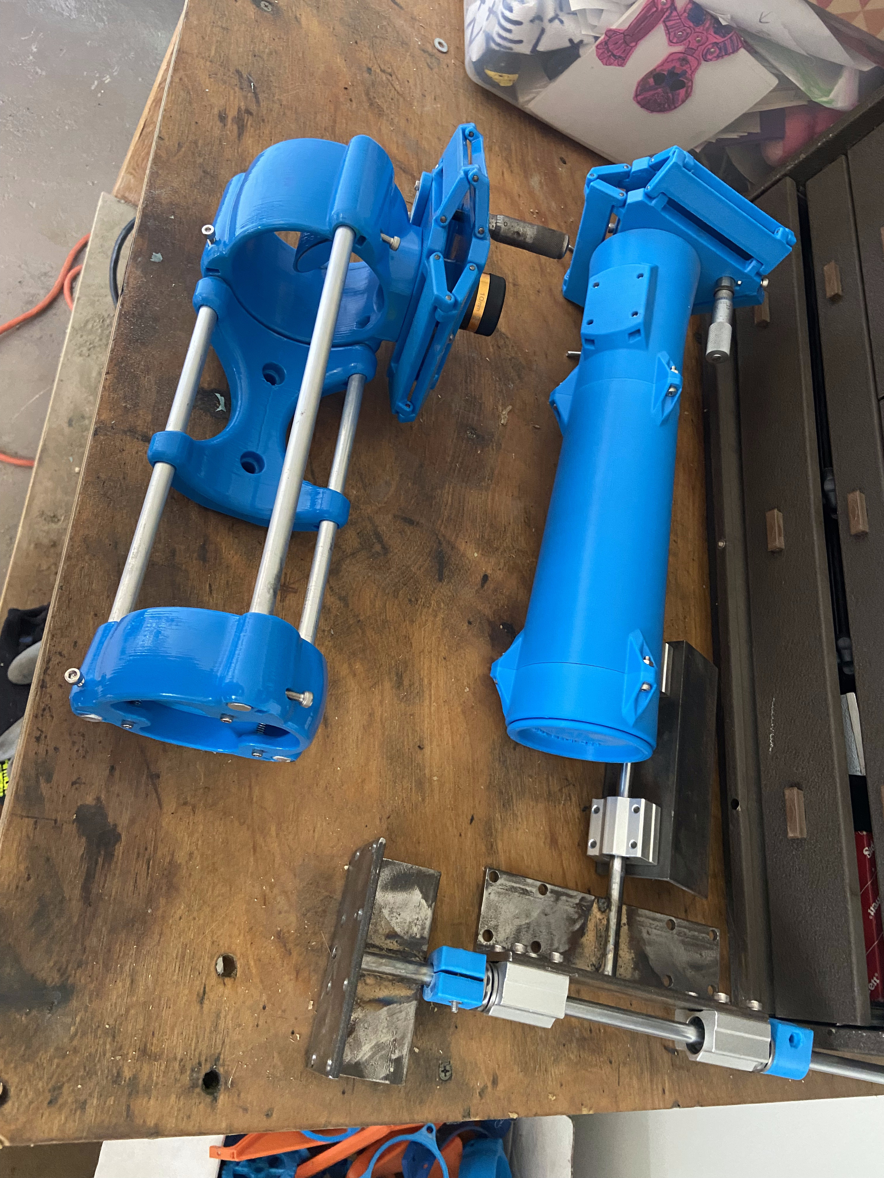 Equatorial mount axis housings disassembled on a workbench — blue printed parts, aluminium rod, worm gear components